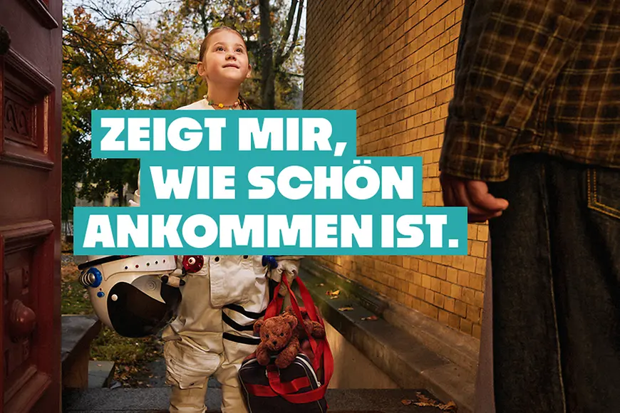 Ein Kind in einem Astronautenanzug steht vor einer geöffneten Haustür, darüber steht der Satz "Zeig mir, wie schön Ankommen ist"