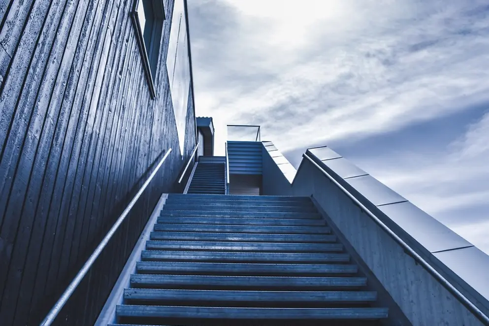 Eine nach oben laufende, steile, breite Treppe mit Handlauf vor einem wolkigen Himmel 