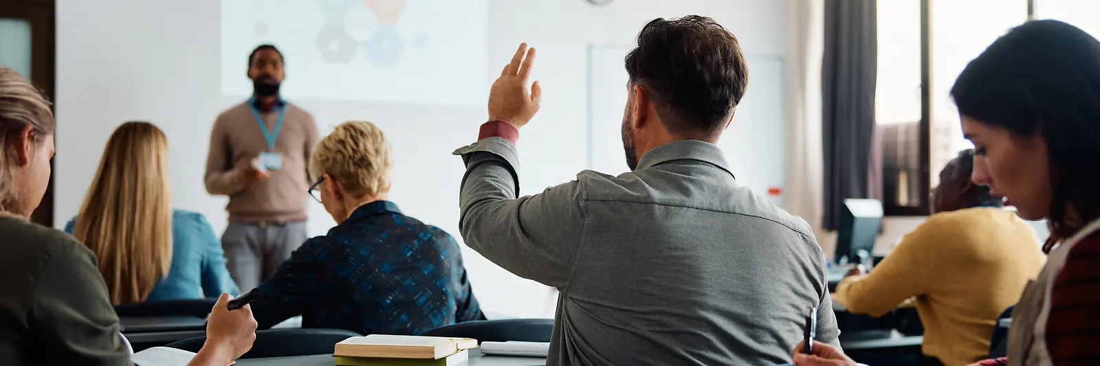 Ein Lehrer erklärt etwas vor einer Gruppe Erwachsener in einem Klassenraum.