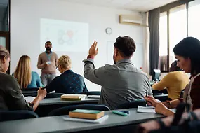 Ein Lehrer erklärt etwas vor einer Gruppe Erwachsener in einem Klassenraum.